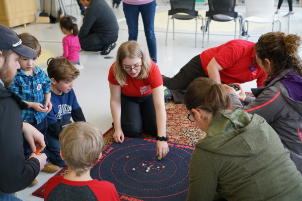 A teen volunteer at The Corning Museum of Glass leads a group children and adults in a game of marbles.